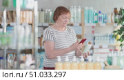 Near display case in pharmacy, senior woman scans barcode on hair loss shampoo. Client collects customer information online, seek reviews and recommendations for use. Стоковое видео, видеограф Яков Филимонов / Фотобанк Лори