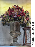 An elegant composition of artificial flowers in a vintage urn vase. The bouquet is dominated by burgundy roses, complemented by green foliage, dried flowers and decorative elements. Next to the vase is an antique lantern, creating an atmosphere of coziness and retro style. Ideal decor for interiors, photo shoots and event design. Стоковое фото, фотограф Анна Кривицкая / Фотобанк Лори
