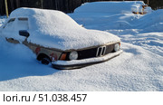 An old car, partially covered in snow, sits in an open area in winter. The car is abandoned and buried in deep snow, and its bodywork, with visible rust spots, indicates a long period of inactivity. In the background, snow-covered logs and a dark forest create an atmosphere of cold and silence. (2021 год). Редакционное фото, фотограф Анна Кривицкая / Фотобанк Лори