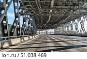 A view from inside a metal bridge in St. Petersburg with its characteristic truss structures and diagonal braces. The sun's rays create a contrasting pattern of shadows on the road surface. In the distance, city buildings and 110 km/h speed limit signs are visible, giving the frame dynamism and urban rhythm. Стоковое фото, фотограф Анна Кривицкая / Фотобанк Лори