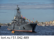 Russian Navy warship No. 130 is anchored in the Neva River in St. Petersburg. The vessel is decorated with flag colors of signal flags stretching from the mast to the bow and stern, which gives it a festive and solemn look. The Russian naval ensign flies from the mast, and the St. Andrew's flag flies from the stern. In the background, the historic buildings and golden domes of St. Petersburg's churches are visible, creating a contrast between the modern warship and the architectural heritage of the city. Clear weather, blue skies and calm water emphasize the atmosphere of a solemn maritime event, probably related to a parade or a Navy holiday. (2015 год). Редакционное фото, фотограф Анна Кривицкая / Фотобанк Лори