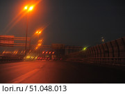 Night view of the Saint Petersburg Ring Road, illuminated by bright street lamps with a warm orange light. The road surface sparkles in the light of headlights and lanterns, creating an atmosphere of movement in the evening city. In the foreground are several cars moving in the indicated directions: Vyborgskoye Highway, Primorskoye Highway, Kronstadt. High noise protection screens are installed on the right, and information signs and signs are above the road. The scene conveys a sense of urban dynamics, high-speed traffic and infrastructure of a megalopolis in the dark. Стоковое фото, фотограф Анна Кривицкая / Фотобанк Лори