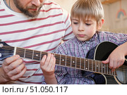 Handsome dad teaches his cute son to play the guitar sitting on sofa in the living room at home. Happy Father's Day. Father teaching his child to play guitar. Kid Learning music skill. Art education. Стоковое фото, фотограф Марина Демидюк / Фотобанк Лори