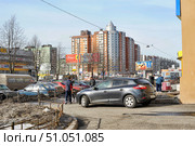 An urban street landscape on a clear sunny day. In the foreground is a parked dark gray hatchback and several people, one of whom is standing next to the car, and the other is walking towards the parking lot. On the left are rows of parked cars and a section of fencing separating the pedestrian zone from the roadway. In the background are modern multi-story residential buildings with red brick and gray metal facades, as well as older panel houses in green and gray. Billboards and store signs, including a supermarket and an optician, are installed along the street. On the sidewalk on the right, several people are busy with their own affairs. In the foreground, the remains of dirty snow are noticeable, typical for late winter or early spring. (2013 год). Редакционное фото, фотограф Анна Кривицкая / Фотобанк Лори