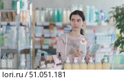 Young woman in sweater standing near racks of medicines and pills and choosing moisturizer with collagen and body lotion at pharmacy. Стоковое видео, видеограф Яков Филимонов / Фотобанк Лори