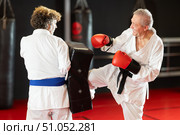 Elderly man in white kimono kicks and punches a punching bag held by trainer. Стоковое фото, фотограф Яков Филимонов / Фотобанк Лори