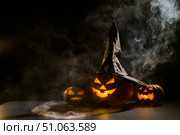 Three creepy Halloween grinning pumpkins glow in the dark among the fog. jack-o-lantern in a witch hat on a black background in smoke. Стоковое фото, фотограф Михаил Решетников / Фотобанк Лори
