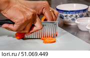 Hands using a crinkle cutter to slice fresh orange carrot rounds on a light cutting board, with patterned bowls and small white dishes in the blurred background, suggesting preparation of fresh ingredients for cooking. Стоковое фото, фотограф Ксения Чунаева / Фотобанк Лори