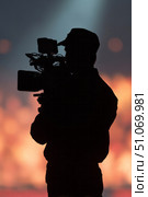 A silhouette of a camera operator is seen holding a professional video camera against a dramatic blurred background featuring bokeh lights and an intense warm glow, possibly from fire or stage lights. The figure is sharply outlined, wearing a cap and uniform, suggesting a production environment such as a live event, film set, or news coverage. The focus on the silhouette creates a cinematic and mysterious mood, emphasizing the act of filming rather than the identity of the person. Стоковое AI-изображение, AI-художник Ксения Чунаева / Фотобанк Лори