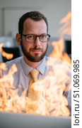A man in a formal office environment, wearing a light blue dress shirt, a patterned yellow tie, and black-framed glasses. He has short dark hair, a neatly groomed beard, and an intense, focused expression. In front of him, large flames dominate the foreground, partially obscuring his body and suggesting either a literal fire or a metaphorical representation of chaos, urgency, or high-pressure situations. The background is softly blurred, keeping the attention on the subject and the dramatic flames, which appear vivid and dynamic, casting a warm glow on his face. The contrast between his composed demeanor and the chaotic element of the fire creates a visually striking and somewhat ironic scene. Стоковое AI-изображение, AI-художник Ксения Чунаева / Фотобанк Лори