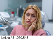 Красивая молодая женщина сидит в современном стоматологическом кабинете, приложив руку к щеке от зубной боли. Стоковое AI-изображение, AI-художник Максим Коломыченко / Фотобанк Лори