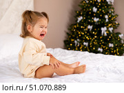 Sad and depressed toddler girl in casual bodysuit sits on a bed in front of a blurred Christmas tree background and crying. Childhood and emotions concept. Стоковое фото, фотограф Ирина Михайличенко / Фотобанк Лори
