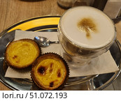 A delicious serving of latte coffee with thick milk foam and caramel pattern on the surface in a double glass mug. Next to it, on a metal tray, are two traditional Portuguese pastel de nata tartlets with a golden caramelized crust and delicate cream inside. Dessert and coffee are served on a white paper napkin with a small teaspoon. The warm wooden table and soft lighting create an atmosphere of coziness and relaxation, ideal for a coffee shop or home tea party. Стоковое фото, фотограф Анна Кривицкая / Фотобанк Лори