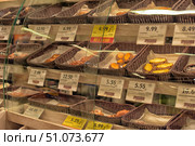 A display case with baked goods in a supermarket, divided by glass partitions. On the shelves are wicker baskets with various types of baked goods, pies and cookies. Each basket has a price tag indicating the cost per piece or kilogram. Among the assortment, you can see traditional products such as Karelian pies, cookies with curd filling and puff pastries. The price tags are in Estonian, and some products are highlighted with yellow price tags with discounts. The lighting is warm, emphasizing the golden color of the baked goods and creating a cozy atmosphere. (2015 год). Редакционное фото, фотограф Анна Кривицкая / Фотобанк Лори