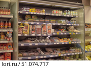 A refrigerated display case in a supermarket with a variety of products. The shelves are lined with sausages, ham, bacon, sliced meat, cheeses, pates, canned goods and sauces. At the top of the display case are jars of sauces, mustard and yoghurts, below are packages of vacuum-packed meat products and cheese slices. On the adjacent counter on the left are bottles of sauces and packages of seasonings, on the right is a basket of baked goods. The products are neatly laid out, the price tags are clearly visible, creating the atmosphere of a store with a wide selection of fresh products. (2015 год). Редакционное фото, фотограф Анна Кривицкая / Фотобанк Лори