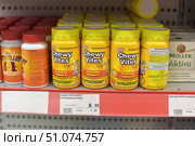 A vitamin store shelf displaying Chewy Vites Kids Vitamin D chewable vitamins. The jars are bright yellow with yellow lids and a colorful label featuring a cartoon character. The front states that the product contains vitamin D, which is good for bone health and immunity, and does not contain artificial flavors. Each package contains 60 chewable tablets with natural fruit flavors. In the foreground is a price tag with a price of 8.95 euros. On the left are other vitamin jars, on the right is a package of Möller Aktiva. The lighting in the store is bright, the photo is taken with the focus on the central packages. (2015 год). Редакционное фото, фотограф Анна Кривицкая / Фотобанк Лори