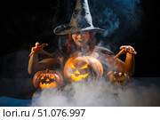 An ominous witch in a hat conjures over a jack-o-lantern. Traditional halloween characters. Mystical fog creeps over pumpkins with carved terrible faces. Woman in carnival costume. Стоковое фото, фотограф Михаил Решетников / Фотобанк Лори