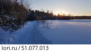 A winter landscape with a snow-covered path leading through the forest, taken at sunset. The sun is low on the horizon, golden rays break through the bare branches of the trees, painting the snow in warm shades. On the left are dense coniferous and deciduous trees covered in snow, on the right is a spacious frozen field or lake, completely covered in untouched snow cover. The sky is clear, with a slight blue tint, creating an atmosphere of peace and quiet. Стоковое фото, фотограф Анна Кривицкая / Фотобанк Лори