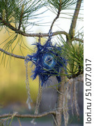 A handmade pendant in the form of a woven amulet or talisman made of blue and light blue threads. The product has the shape of a star with fluffy fibrous edges and is attached to a coniferous tree branch with a rope. The background shows softly blurred green and yellow shades of nature, creating an atmosphere of warmth and comfort. The amulet looks like an ethnic or handmade element, harmoniously integrated into the natural environment. Стоковое фото, фотограф Анна Кривицкая / Фотобанк Лори