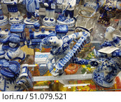 A display case with a collection of porcelain figurines in the traditional Russian Gzhel style. Each piece is hand-painted in characteristic blue and white tones with floral and geometric patterns. In the center of the composition is a figurine of an accordionist in a dynamic pose, holding an accordion and with a pronounced facial expression. Nearby are miniatures of a steam locomotive, figurines of people in folk costumes and scenes from village life. In the background, crystal glasses with cup holders and other elements of Russian decorative tableware are visible. The bright lighting of the display case emphasizes the shine of the glaze and the richness of the cobalt shades. Стоковое фото, фотограф Анна Кривицкая / Фотобанк Лори