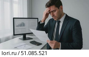A young Caucasian man in a suit looks stressed while holding a document. He stands near a desk with a computer displaying graphs and a calculator. Стоковое AI-видео, аниматор Nart / Фотобанк Лори