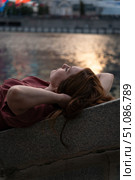 Young woman with red hair in a burgundy dress relaxing by the riverbank at sunset, with warm golden reflections shimmering on the water. Стоковое фото, фотограф Софья Соломонова / Фотобанк Лори