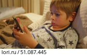 Cute kid boy in pajamas lying in bed watching smartphone, smiling. Children's screen time. Child plays games using phone at the evening or night. Social media addiction. Childhood, modern technology. Стоковое видео, видеограф Марина Демидюк / Фотобанк Лори