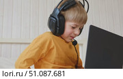 Little smiling 6 years old boy using a laptop with headphones. Child having video call with friends, granny at home in bed. Kid looking at display and waving hand. Childhood leisure activity concept. Стоковое видео, видеограф Марина Демидюк / Фотобанк Лори