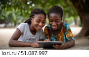 Two African girls sharing a tablet to study together outside under a tree in an educational environment. Black African children from disadvantaged families study in school, read books, receive primary education, volunteer service. Стоковое AI-изображение, AI-художник ALLA YURTAYEVA / Фотобанк Лори