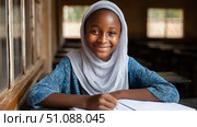 Young smiling African girl in hijab sitting near the window and studying in an African school. Lack of school supplies. Black African children from disadvantaged families learn in school, read books, get primary education, volunteer service. Стоковое AI-изображение, AI-художник ALLA YURTAYEVA / Фотобанк Лори