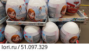A photo of a store display case with large plastic surprise eggs featuring characters from the Disney cartoon Frozen. The packaging features Anna, Elsa, and the snowman Olaf on a blue and pink background with icy patterns. The eggs contain marmalade and a board game for the whole family, as indicated on the packaging. The inscription Frozen is visible in the center, and the Disney logo is on the side. In the background, you can see eggs featuring characters from the cartoon Cars. The bright and colorful children's products are presented in boxes and are intended as gifts for children. (2018 год). Редакционное фото, фотограф Анна Кривицкая / Фотобанк Лори