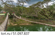 Two rickety Bamboo Hanging Bridges over a tropical river. The weaving of paths, unreliable railings is visible. Houses in the distance, among the green vegetation. Clouds in the sky. Philippines.Bohol (2024 год). Стоковое фото, фотограф Вера Ивановна Кубарева / Фотобанк Лори