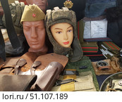 A display case with military paraphernalia and equipment. In the foreground are mannequins with headgear: a cap with a red star and a tank helmet with a camouflage pattern. Nearby are brown leather holsters, army belts, shoulder straps of different colors and ranks, as well as a compass and various accessories. In the background are books with the inscription "Instructions" and elements of a military uniform. The photo conveys the atmosphere of a military store, museum or collector's market, where items of army equipment and Soviet symbols are presented. Стоковое фото, фотограф Анна Кривицкая / Фотобанк Лори