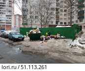 A photograph of a city courtyard with a garbage container site. In the foreground is an overflowing green garbage container, around which cardboard boxes, bags and household waste are scattered. Next to it stands a man in dark clothes and a green vest, holding a yellow bag. In the background are multi-story Soviet-era apartment buildings with balconies and windows. The site is covered with the remains of dirty snow, and the road is covered with puddles and ice. The atmosphere emphasizes the problems of landscaping, overflowing garbage containers and untimely waste removal. The scene reflects a typical winter urban landscape of Russian courtyards. (2019 год). Редакционное фото, фотограф Анна Кривицкая / Фотобанк Лори