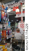 The photo was taken in a checkout line at a large shopping center. The frame shows the checkout area with a salesperson, customers, and a counter with impulse goods. The display case contains packages of condoms from well-known brands Durex and Contex, as well as related small goods - toothbrushes, batteries, patches. In the background, the sign of the Maksidom store and the checkout number 7 are visible. Above the checkouts, there is a festive decoration - a white garland with red balls. The information signs indicate payment methods, including Visa and MasterCard bank cards. The atmosphere of everyday shopping and excitement in a department store. (2018 год). Редакционное фото, фотограф Анна Кривицкая / Фотобанк Лори