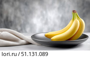 Fresh bananas arranged on a black plate with a soft cloth on a table. Стоковое AI-изображение, AI-художник Katsiaryna Repich / Фотобанк Лори