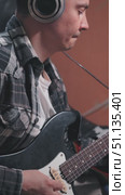 A musician wearing large headphones is focused on playing an electric guitar. The plaid shirt adds a casual vibe. Close-up of playing an electric guitar in a home studio. Стоковое видео, видеограф Воловодов Владимир / Фотобанк Лори