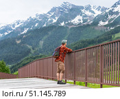 Male hiker in red plaid shirt and backpack stands on wooden bridge, gazing at majestic snow-capped mountains, surrounded by lush greenery, enjoying nature's beauty and tranquility, hiking and tourism. Стоковое фото, фотограф Елена Гоосен / Фотобанк Лори