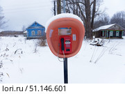 A winter rural landscape with a street payphone in the center of the frame. The red phone is installed in an orange protective booth, covered with snow on top. In the background, two wooden village houses are visible - one is blue, the other is dark with green elements. There is white fluffy snow around, dry grass sticking out from under the snowdrifts is noticeable. The atmosphere of solitude and provincial Russia is emphasized by the contrast of old houses and a street payphone, rare for today. Стоковое фото, фотограф Анна Кривицкая / Фотобанк Лори