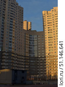 A modern multi-story residential building in the rays of the sunset. The high-rise building made of light brick with numerous windows and glazed loggias reflects the warm sunlight. The architecture is typical for urban areas: monolithic brick construction, strict geometric lines and high floors. In the foreground you can see a street lamp, a fence and small buildings that create an atmosphere of an urban environment. The image is suitable for illustrating topics related to urbanism, architecture, construction, residential areas and modern city life. (2015 год). Стоковое фото, фотограф Анна Кривицкая / Фотобанк Лори