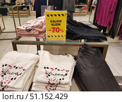 The photo was taken in a clothing store. In the foreground, on a glass table, women's white Guess T-shirts with an original print in the form of cosmetics and an inscription on the chest, as well as gray and black jeans are neatly laid out. In the background, folded pink T-shirts are visible. In the center of the table, there is a bright yellow sign with the inscription "Calvin Klein -70%" on a red background, framed by a black and yellow stripe, which attracts the attention of buyers to the sale. Part of the sales area is visible in the frame: hangers and racks with other clothes, creating the atmosphere of a modern boutique. (2019 год). Редакционное фото, фотограф Анна Кривицкая / Фотобанк Лори