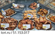 Display cases in a supermarket with ready-made meat dishes. In the foreground are trays with baked and fried chicken legs, thighs and wings, seasoned with spices and having a golden-brown crust. In the center are skewers with shashlik from chicken fillet, marinated and prepared for the grill. In the background are trays with grilled chicken halves and other meat products. Above the dishes are price tags indicating the cost per kilogram, composition and promotional offers. The photo conveys the atmosphere of the grocery department of the supermarket, the appetizing appearance of the dishes and the convenience of buying ready-made food. Suitable for illustrating culinary publications, online store catalogs, articles on nutrition and ready-made solutions for dinner. Редакционное фото, фотограф Анна Кривицкая / Фотобанк Лори