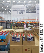 Inside a large wholesale store, goods are laid out on shelves and pallets. In the foreground are cardboard boxes with gas cylinders for the Sledopyt camping stoves and canisters with liquid in plastic bottles. A large price tag hangs above the goods indicating the cost of 149.99 rubles for a set of two gas cylinders. In the background, customers are visible, metal racks with boxes and bright industrial lighting. The scene conveys the atmosphere of a warehouse hypermarket where goods for the country, hiking and everyday needs are sold. (2019 год). Редакционное фото, фотограф Анна Кривицкая / Фотобанк Лори