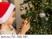 Close up side view of a cute happy little boy in red Santa hat decorating a Christmas tree in the living room at home. Merry Christmas and happy New Year winter holidays. High-quality photo. Стоковое фото, фотограф Ирина Михайличенко / Фотобанк Лори