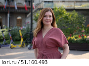 Confident middle-aged woman in a rose jumpsuit stands outdoors with colorful flowers and e-scooters in the background. Стоковое фото, фотограф Софья Соломонова / Фотобанк Лори