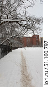 Winter cityscape. In the foreground, a path is visible, trodden in deep snow, leading along a metal fence and into the distance to residential multi-story red brick buildings. On the left, spreading trees with bare branches covered with a fresh layer of snow create the effect of an arch over the path. In the background, benches are visible, buried in snow, and city buildings. The scene conveys the atmosphere of a quiet winter day, the calm and comfort of an urban winter. Стоковое фото, фотограф Анна Кривицкая / Фотобанк Лори