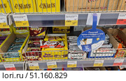 Supermarket display cases with chocolate bars and sweets from various brands. In the foreground, you can see brightly colored packages of Nuts, Twix, Kinder Bueno, Fun Banan, Candy Nut, Marzipan and other bars. All the products are neatly arranged in boxes with price tags indicating the cost and weight. The display case is made in a standard self-service store, with metal shelves and dividers. In the center of the photo, there is an advertising sign offering Moomin stickers and tattoos for purchases over a certain amount. The bright composition emphasizes the variety of products and the atmosphere of a grocery store. (2021 год). Редакционное фото, фотограф Анна Кривицкая / Фотобанк Лори