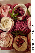 A set of elegant sweets in the form of flowers. Desserts are made in the form of roses and peonies from protein or butter cream, painted in delicate shades of white, pink and burgundy. The petals look realistic and are decorated with small silver sugar beads, which gives the composition sophistication and a festive mood. Sweets are wrapped in burgundy paper, reminiscent of the design of a flower bouquet, and complemented by green decorative leaves. The photo conveys the atmosphere of celebration and is ideal for illustrating confectionery, holiday gifts and ideas for decorating desserts. Стоковое фото, фотограф Анна Кривицкая / Фотобанк Лори