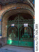 Entrance arch with decorative green metal gates in a historic building in St. Petersburg. The gates are made in the style of artistic forging with floral ornaments and patterns in the form of curls and rosettes. The yellow brick arch is decorated with colored ceramic tiles with geometric and floral ornaments, creating an Art Nouveau atmosphere. Behind the gates, you can see a passage into a courtyard-well, typical of the architecture of St. Petersburg. On the gates there are information signs and a warning sign about the work of a tow truck. The atmospheric photo reflects the beauty of the city's architecture and the details of old houses. (2019 год). Редакционное фото, фотограф Анна Кривицкая / Фотобанк Лори