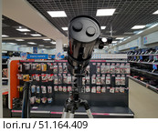 Electronics and equipment store. In the foreground, a close-up of a telescope on a tripod with a protective cover is shown, installed in the sales area. The optics and mounts are visible, which emphasizes the technical details of the device. In the background are display cases with accessories: memory cards, USB drives, adapters and other equipment. You can also see shelves with laptops and other equipment. The bright lighting of the store emphasizes the details, creating the atmosphere of a modern retail space with a wide selection of goods. The photo demonstrates the combination of astronomical equipment and electronics in retail. (2019 год). Редакционное фото, фотограф Анна Кривицкая / Фотобанк Лори